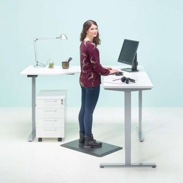 Nainen seisomassa Economy Flex -sähköpöytä, 3-jalkainen L-malli seisomapöydän edessä.