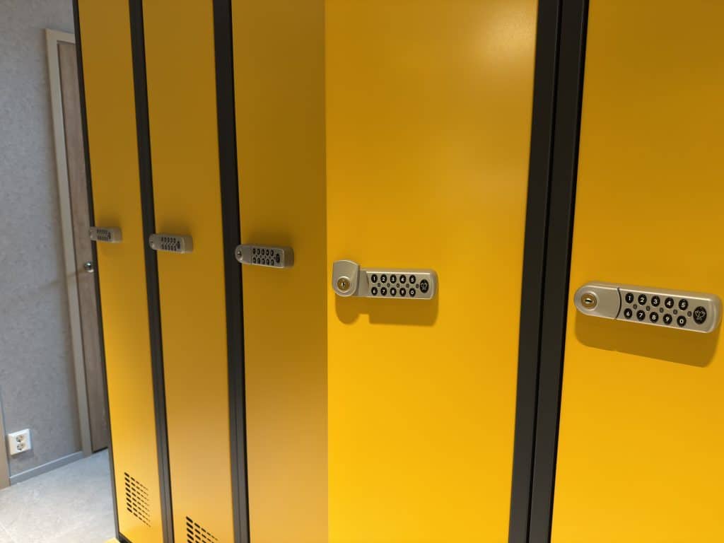 Rivi keltaisia ilmastoidut pukukaapit, joissa on mustat reunat ja elektroniset yhdistelmälukot, jotka sijaitsevat sisätiloissa logistiikkakeskuksessa vaalean seinän ja oven vieressä.